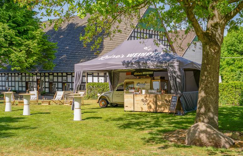 Luises Weinbar - Exklusives Catering-Zelt in Bielefeld vor Fachwerkhof Ein graues Mastertent-Catering-Zelt namens "Luises Weinbar" vom Weinparadies Hess in Bielefeld, auf einer grünen Wiese mit Stehtischen und einem Foodtruck, vor einem historischen Fachwerkhof.