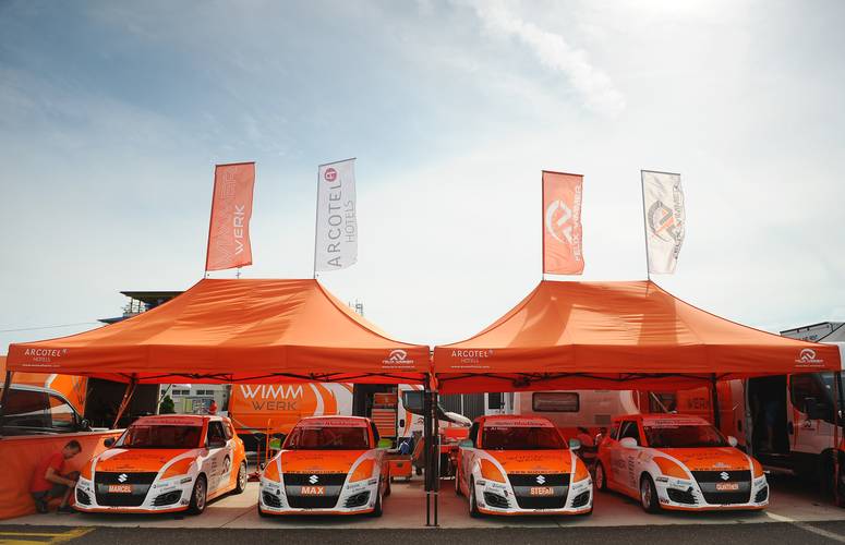 Mastertent Eventzelte für Motorsport-Teams: Wimmer Werk Racing im Rennsport-Paddock Professionelles Wimmer Werk Racing Team Paddock-Setup mit zwei orangefarbenen Mastertent Faltpavillons und mehreren Werbe-Fahnen im Wind. Vier Suzuki Rennwagen mit Fahrer-Namen sind unter den Zelten aufgereiht.